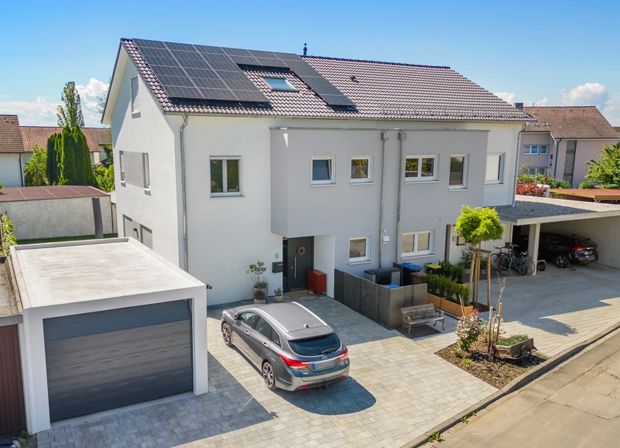 Moderne Doppelhaushälfte mit Solaranlage auf dem Dach, grauem Auto und Garage, klarer Himmel im Hintergrund.