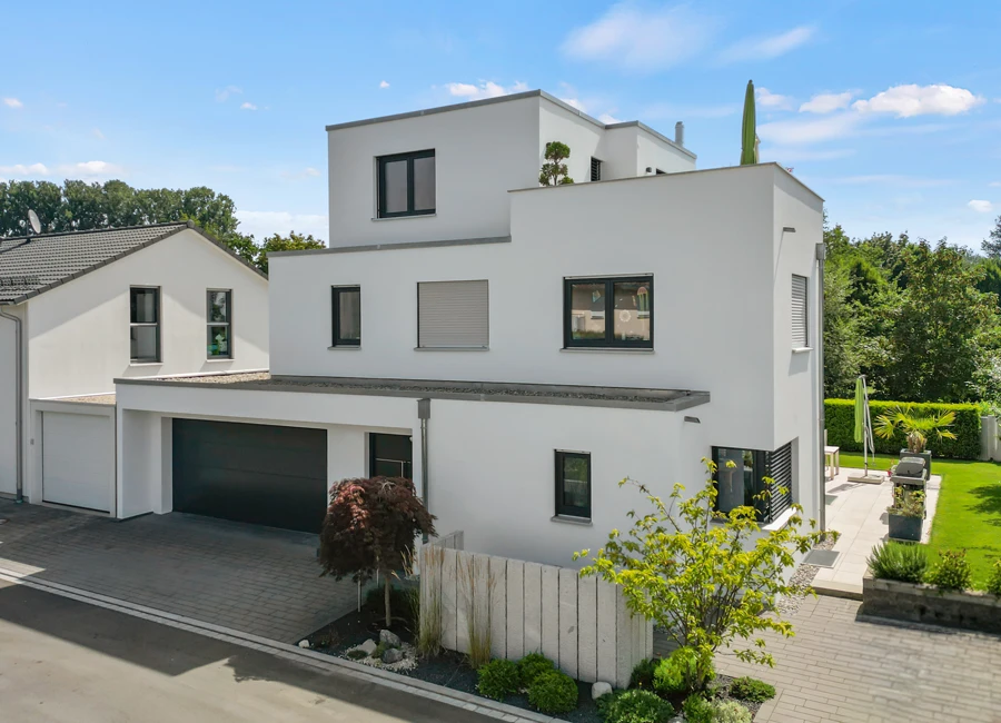 Modernes weißes Haus mit Flachdach und Garten, umgeben von Bäumen an einem sonnigen Tag, ruhige Wohnumgebung.