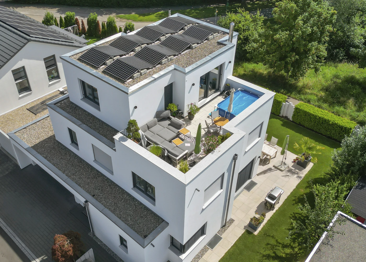Modernes Haus mit Solaranlagen, Dachterrasse, Pool und Garten, umgeben von grüner Landschaft, aus der Vogelperspektive.