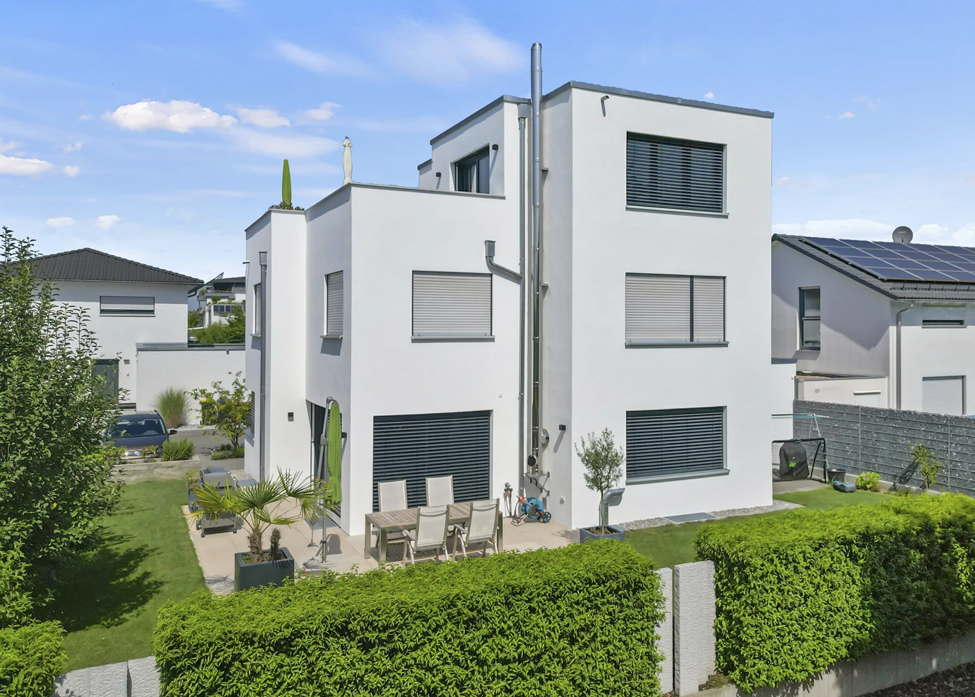Modernes weißes Wohnhaus mit Terrassenbereich und Garten, umgeben von Hecken und angrenzend an andere Häuser.
