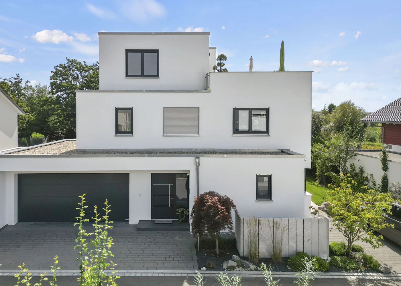 Modernes kubusförmiges Einfamilienhaus mit Flachdach und großen Fenstern, umgeben von Garten und blauen Himmel.