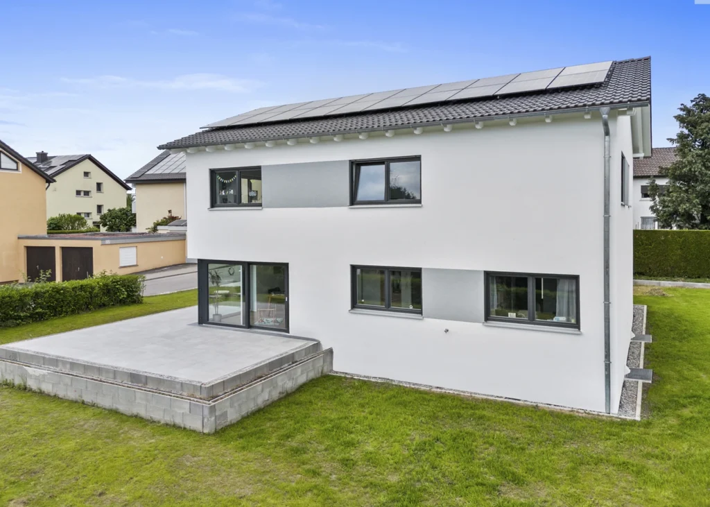 Modernes Einfamilienhaus mit Solarpanelen auf dem Dach und großer Terrasse, umgeben von grünem Garten.