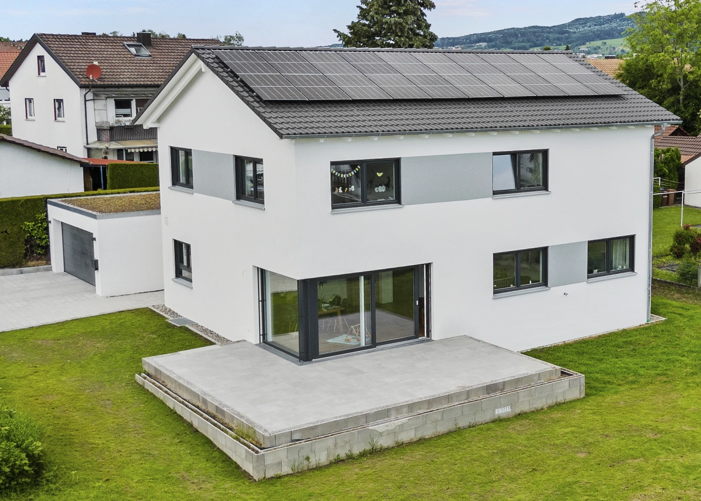 Modernes Einfamilienhaus mit Solarmodulen auf dem Dach, umgeben von grünem Garten und Nachbarhäusern.