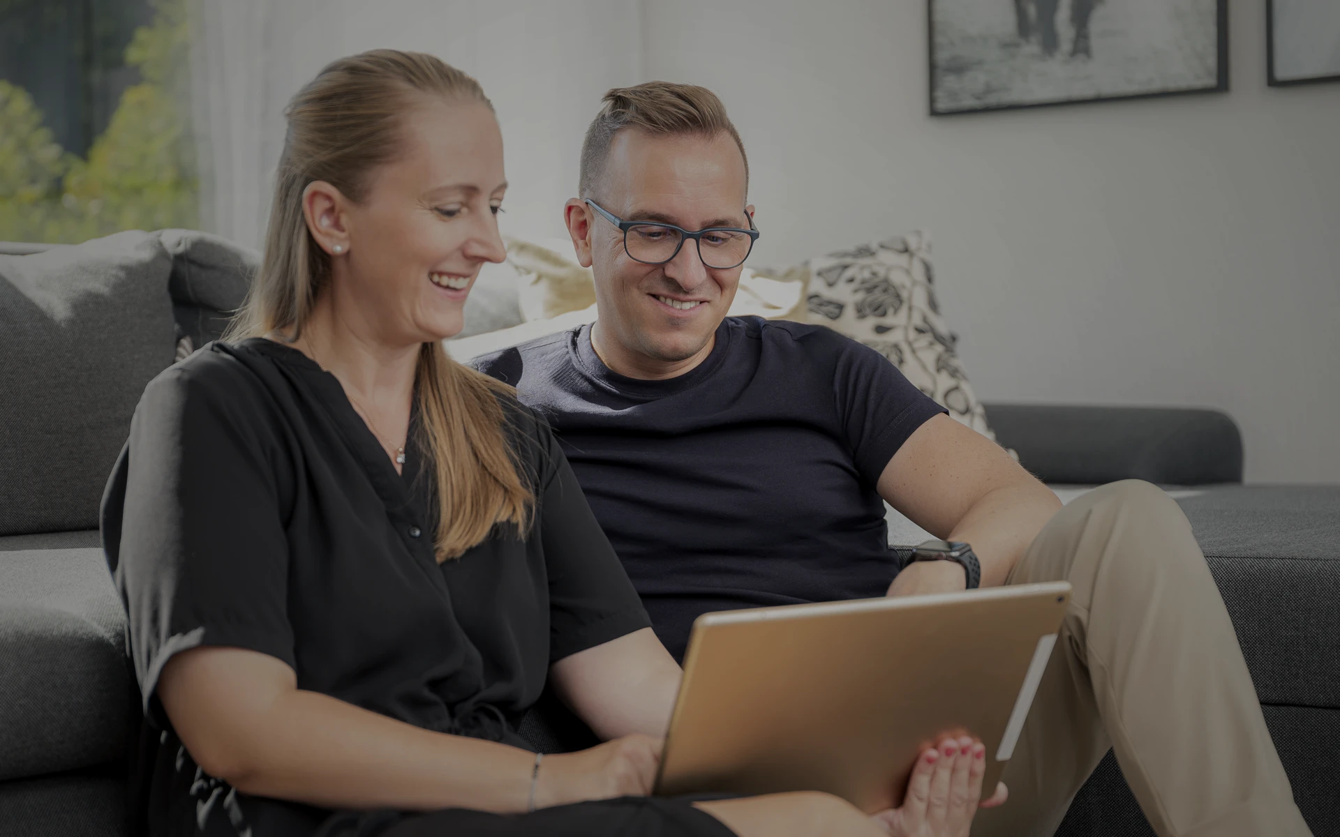 Ehepaar sitzt vor der Couch am Boden und schauen sich eine Webseite auf einem Tablet an.