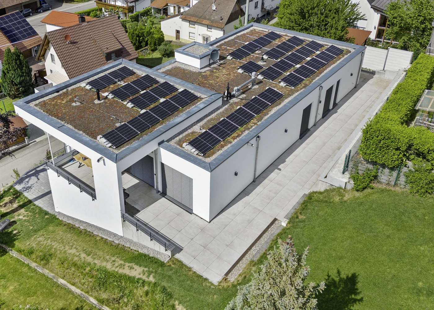 Flachdach mit Solaranlagen auf modernem Gebäude in Wohngebiet, umgeben von grünen Gärten und traditionellen Häusern.